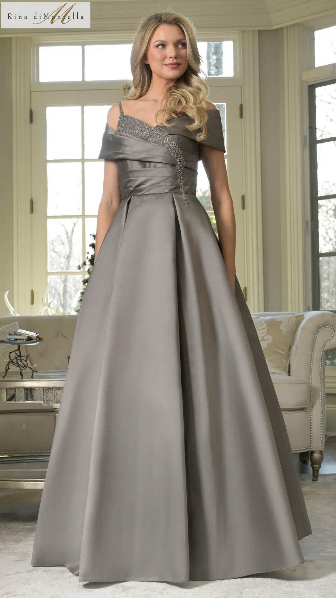 A woman in a gunmetal Long Formal Dress: Rina Di Montella RD3221 with an off-shoulder neckline stands in a bright living room with large windows.